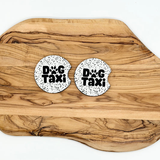 Two round white coasters with black speckled texture and the words 'DOG Taxi' printed in black, displayed on a wooden cutting board over a white background.