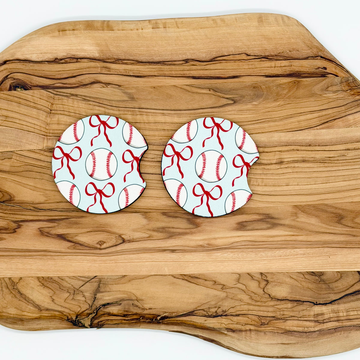 Two round coasters with bite-sized tabs, featuring a white and red baseball pattern with red bows, displayed on a rustic olive wood serving board.